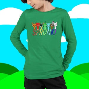 Puede incluir: Una camiseta verde de manga larga con la palabra "SPRUNKI" en letras coloridas, con una fila de personajes de dibujos animados encima. La camiseta est&aacute; hecha de un material suave.