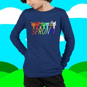 Puede incluir: Una camiseta de manga larga azul marino con la palabra "SPRUNKI" en colores arco&iacute;ris y gr&aacute;ficos de personajes de dibujos animados encima del texto. La camiseta est&aacute; hecha de un material suave.