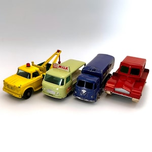 Pode incluir: Quatro caminhões de brinquedo vintage em amarelo, verde, azul e vermelho. O caminhão amarelo é um guincho com uma luz vermelha. O caminhão verde tem um sinal de "Drink More Milk". O caminhão azul é um caminhão-tanque e o vermelho tem uma plataforma.
