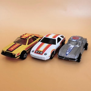 May include: Three toy cars are displayed on a light orange surface. The cars are a yellow sports car with a red and orange design, a white car with red and orange stripes and the number 928, and a silver car with a blue and white design.