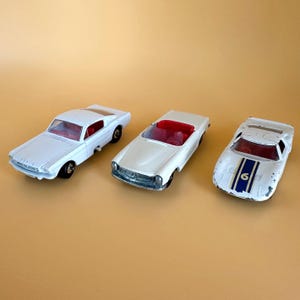 以下が含まれることがあります： 白のヴィンテージおもちゃの車3台。クーペ、コンバーチブル、青いストライプと数字6のレーシングカーが含まれます。車内は赤色で、車輪は黒色です。