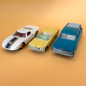 May include: Three die-cast toy cars: a white sports car with a blue racing stripe and the number 6, a yellow convertible with a white interior, and a blue station wagon. Each vehicle features metal bodies, plastic windows, and rolling wheels.