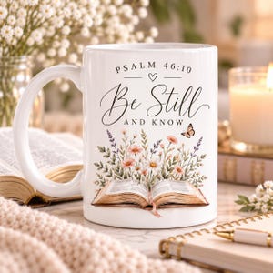 May include: A white ceramic mug featuring the text Psalm 46:10 and Be Still And Know in black script and serif fonts. The design includes a colorful illustration of an open book with a bouquet of wildflowers and a small butterfly above it.