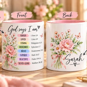 May include: Two white ceramic mugs with pink handles and interiors. The front features a floral bouquet, the text God says I am, and a list of attributes with Bible verses. The back displays a floral bouquet, a pink ribbon, and the name Sarah.