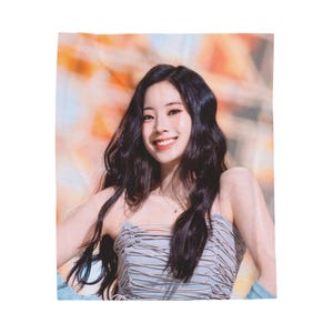 May include: A woman with long black hair is smiling. She is wearing a light blue, ruffled, sleeveless top. The background is a blurred image of a colorful, abstract design.