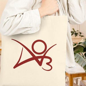 May include: Cream-colored tote bag with a maroon abstract logo. The logo features a circle with curved lines extending outward and a stylized "V" and "3" below. The bag is held by a person wearing a white shirt.