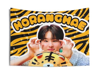May include: A yellow and black striped fabric wall tapestry with the text "HORANGHAE" in large, yellow, bubble letters. A young person with black hair is holding a yellow and orange tiger plush toy in front of their face.