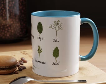 May include: White ceramic mug with a blue interior and handle. The mug features illustrations of herbs like sage, dill, lavender, and mint, with their names printed below. A spoon and a pastry are on a wooden surface.