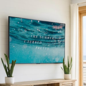 Puede incluir: Un televisor de pantalla plana negro grande montado en una pared blanca muestra el texto "THE SUMMER I TURNED PRETTY" sobre un fondo de agua turquesa. Dos plantas de serpiente en macetas se encuentran en un mueble de madera.