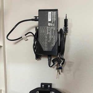 Op de afbeelding: Een zwarte stroomadapter met een label met de tekst "Model: Zac-C02" en verschillende veiligheidscertificeringen. De adapter is aangesloten op een stopcontact en heeft een snoer dat naar beneden hangt. Er hangt ook een set sleutels aan de adapter.