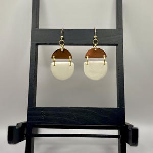May include: A pair of handmade earrings with a geometric design. Each earring features a half-circle of brown material above a white, semi-circular shape. The earrings are suspended from gold-colored hooks.
