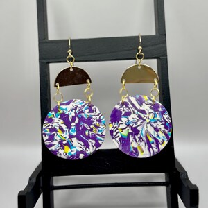 May include: A pair of dangle earrings with a gold-colored half-circle top and a circular bottom. The bottom of the earrings have a marbled design in purple, white, yellow, and blue. The earrings are hanging on a black display stand.
