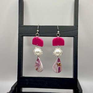May include: A pair of dangle earrings with a vibrant fuchsia top, a pearl-like bead, and a marbled teardrop-shaped bottom. The earrings are displayed on a black stand against a white background. The earrings have silver-colored hooks.
