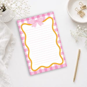 Può includere: Un blocco note rettangolare con uno sfondo a quadretti vichy rosa e bianchi. Il centro presenta un'area di scrittura bianca con linee orizzontali, racchiusa da un bordo arancione ondulato e sormontata da un piccolo fiocco rosa chiaro.