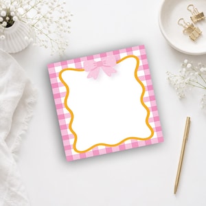 Puede incluir: Un bloc de notas cuadrado con un fondo de patrón de cuadros vichy rosa y blanco. El centro contiene una gran área blanca en blanco enmarcada por un borde dorado ondulado, rematado con una pequeña ilustración de un lazo rosa claro con lunares blancos.