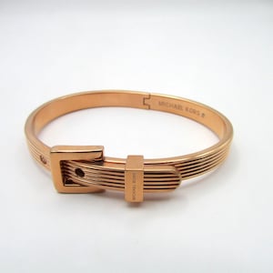 May include: Rose gold-tone metal bracelet with a buckle design. The bracelet features a series of parallel lines and a rectangular buckle closure. The words "MICHAEL KORS" are visible on the bracelet.