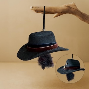 Puede incluir: Un sombrero fedora azul oscuro con ala ancha y una banda de terciopelo rojo oscuro. El sombrero tiene un adorno de plumas decorativas y está suspendido por un cordón negro. Una imagen circular más pequeña del sombrero está en la esquina inferior derecha.