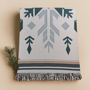 Puede incluir: Una manta tejida plegada con un patrón geométrico en tonos grises, verde azulado y marrón. La manta presenta una franja a lo largo del borde inferior y un diseño decorativo de flechas y diamantes. Una ramita de hoja perenne se coloca en el lateral.