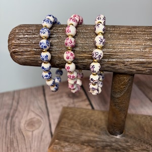 May include: Three beaded bracelets with floral patterns in blue, pink, and purple are displayed on a wooden stand. The bracelets feature white beads with gold accents. The background is a light gray and wooden surface.