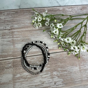 May include: Three beaded bracelets in black, white, and silver. The bracelets are stacked on top of each other on a wooden surface.