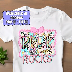 May include: White t-shirt with a colorful graphic that says "PRE-K ROCKS." The design includes a pink bow, flowers, and a pencil. The shirt also has text that says "AVAILABLE IN GRADES PRE-K - FIFTH."