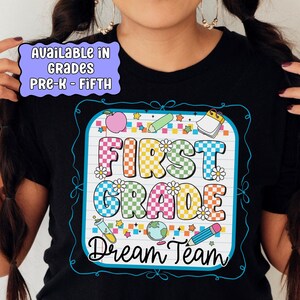 May include: Black t-shirt with a colorful graphic design for First Grade Dream Team. The design includes the words "First Grade" in large, colorful letters, along with school-themed illustrations. The shirt is available in grades Pre-K through Fifth.