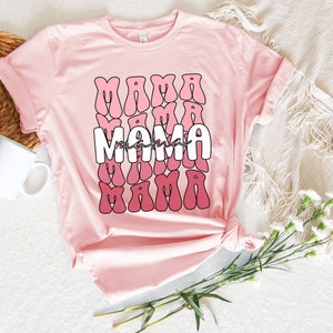 以下が含まれることがあります： ピンクのTシャツに、レトロなスタイルでピンクと白のグラデーションで「ママ」という文字が繰り返しデザインされています。