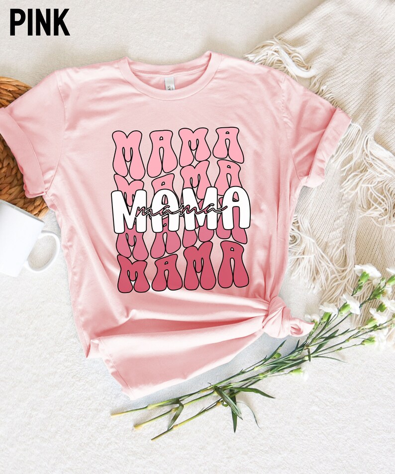 以下が含まれることがあります： ピンクのTシャツに、レトロなスタイルでピンクと白のグラデーションで「ママ」という文字が繰り返しデザインされています。