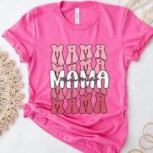 以下が含まれることがあります： ピンクのTシャツに、レトロなスタイルで「ママ」という単語がピンクと赤のグラデーションで繰り返しデザインされています。