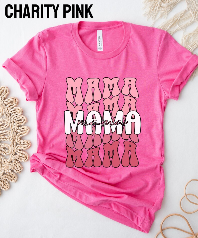 以下が含まれることがあります： ピンクのTシャツに、レトロなスタイルで「ママ」という単語がピンクと赤のグラデーションで繰り返しデザインされています。