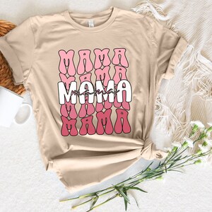 以下が含まれることがあります： タン色のTシャツに、レトロなスタイルで「ママ」という単語が3回繰り返されたグラフィックデザインが施されています。「ママ」という単語は白で、ピンクのアウトラインが描かれています。