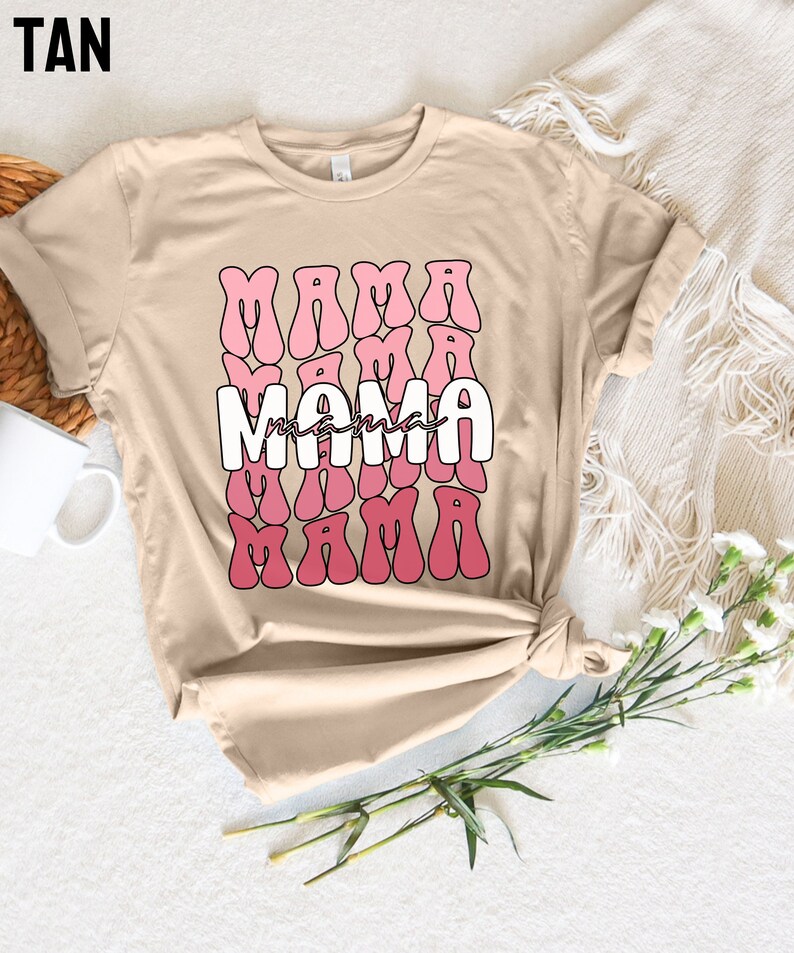 以下が含まれることがあります： タン色のTシャツに、レトロなスタイルで「ママ」という単語が3回繰り返されたグラフィックデザインが施されています。「ママ」という単語は白で、ピンクのアウトラインが描かれています。