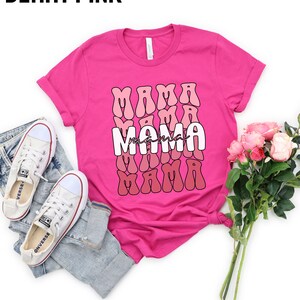 以下が含まれることがあります： レトロなフォントスタイルで「ママ」という単語が繰り返されたピンクのTシャツ。テキストはピンクと赤のグラデーションになっています。