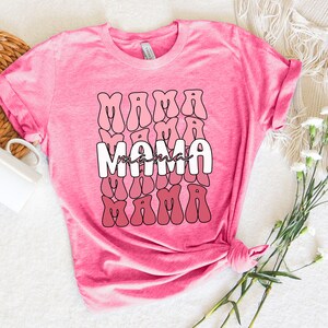 以下が含まれることがあります： ピンクのヘザーTシャツに、レトロなフォントで「mama」と書かれた白いグラフィックがプリントされています。グラフィックは、ピンクのグラデーションで重ねられたパターンで繰り返されています。