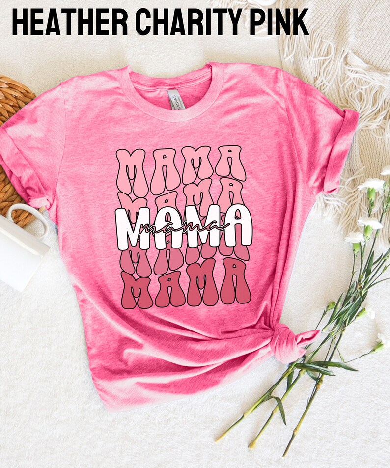 以下が含まれることがあります： ピンクのヘザーTシャツに、レトロなフォントで「mama」と書かれた白いグラフィックがプリントされています。グラフィックは、ピンクのグラデーションで重ねられたパターンで繰り返されています。