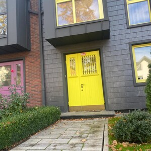 May include: Exterior view of a building with a bright yellow door and matching window frames. The building has gray siding and brick accents. A stone walkway leads to the door, with green bushes and grass on either side.