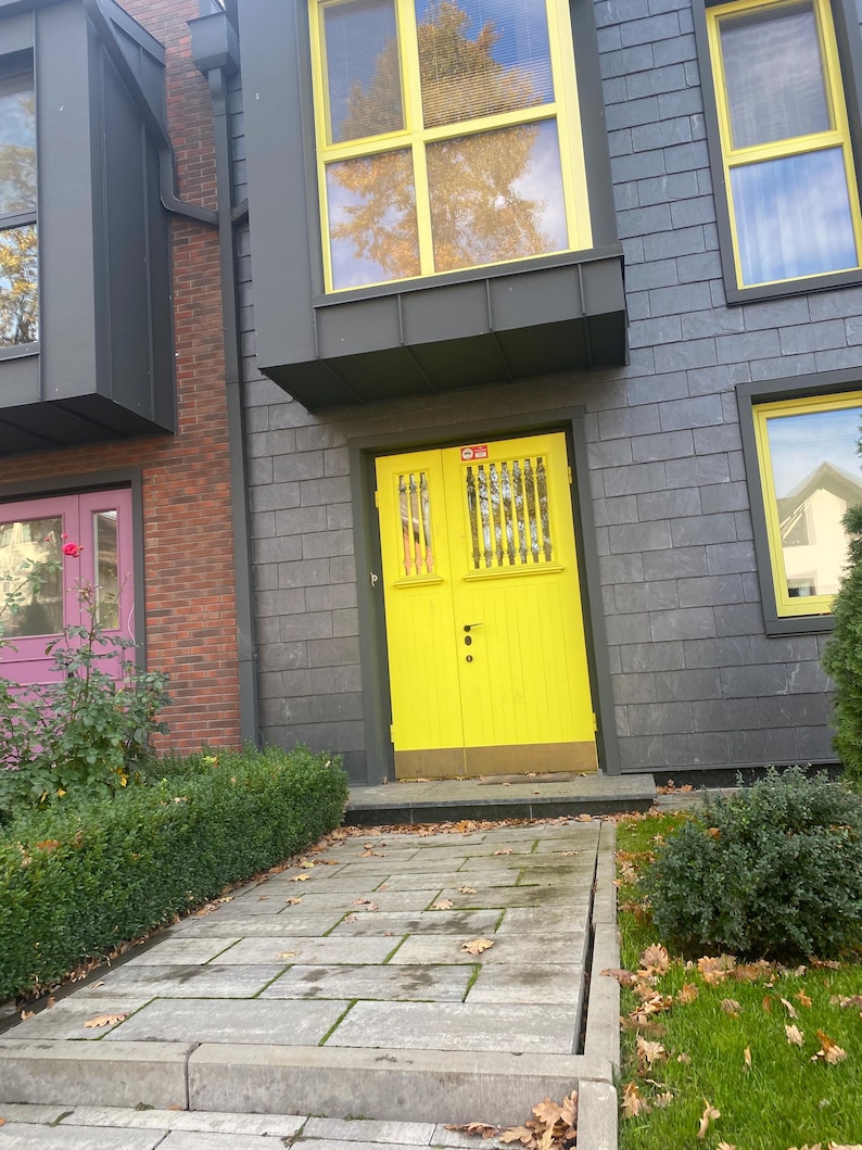 May include: Exterior view of a building with a bright yellow door and matching window frames. The building has gray siding and brick accents. A stone walkway leads to the door, with green bushes and grass on either side.