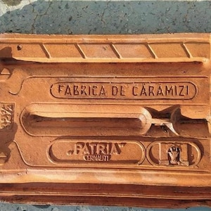 May include: A rectangular, terracotta-colored brick with embossed text and designs. The text includes "FABRICA DE CARAMIZI" and "PATRIA CERNĂUȚI". The brick has a textured surface and is likely a historical building material.