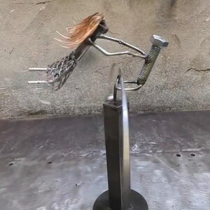 Puede incluir: Escultura metálica de una mujer buceando, elaborada con diversas piezas de metal. La figura tiene cabello color cobre que fluye y está sostenida por un poste vertical sobre una base circular. La escultura se encuentra sobre un fondo neutro.
