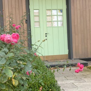 May include: A light green door with a windowed upper section and a smaller window to the left. The door is set in a dark gray frame against a wood-paneled wall. Pink roses and green foliage are in the foreground, with a stone walkway.