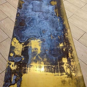 May include: A rectangular table top with a distressed finish. The surface features a blend of gold, navy blue, and black hues, creating a unique, abstract pattern. The gold areas have a metallic sheen, while the blue and black sections appear matte.