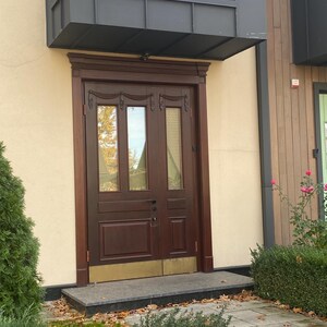 May include: A dark brown wooden double door with glass panels and decorative carvings. The door is set in a light beige wall, with a dark gray awning above. A small stone step leads to the door, with greenery and fallen leaves in the foreground.
