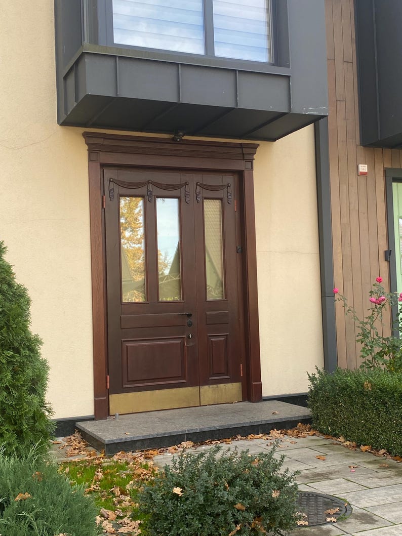 May include: A dark brown wooden double door with glass panels and decorative carvings. The door is set in a light beige wall, with a dark gray awning above. A small stone step leads to the door, with greenery and fallen leaves in the foreground.