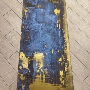 May include: A rectangular, abstract artwork with a distressed appearance. The piece features a combination of gold and dark blue hues, with the blue paint applied in a layered, textured manner over the gold base. The overall effect is a blend of metallic and matte finishes.