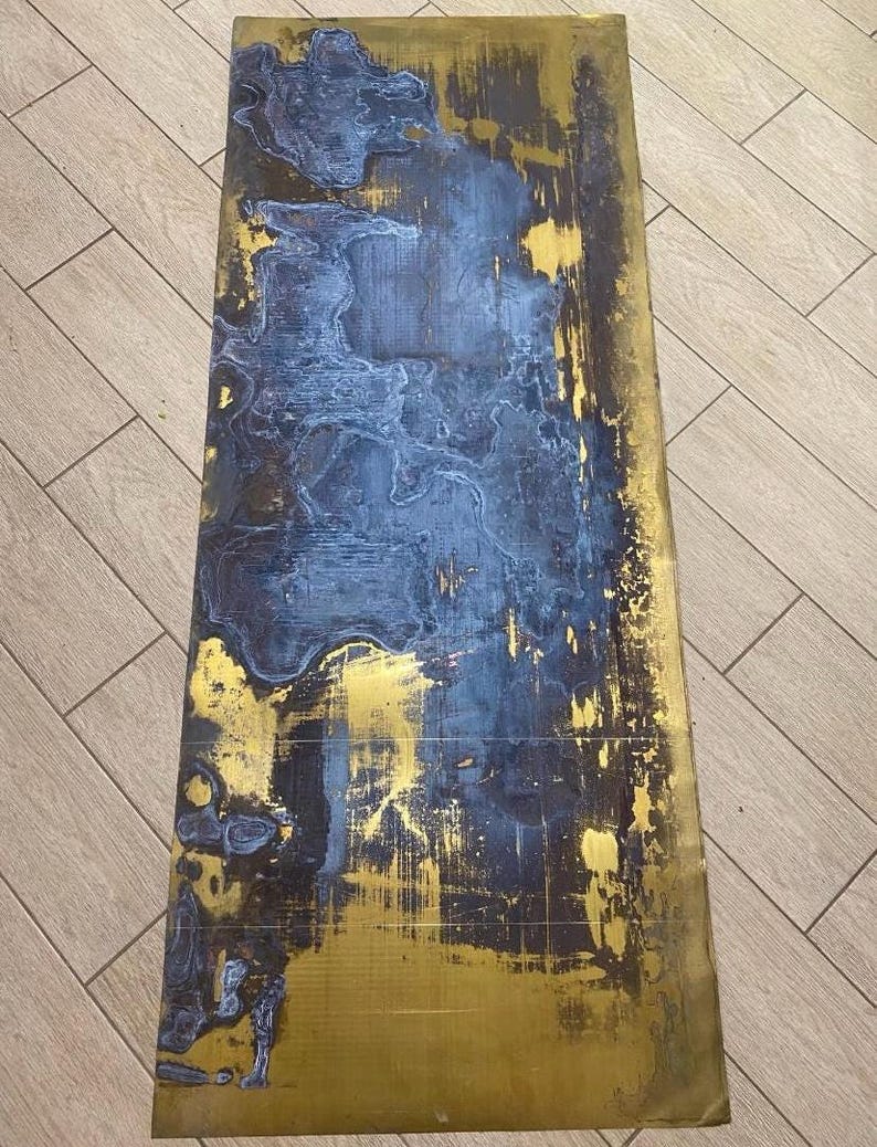 May include: A rectangular, abstract artwork with a distressed appearance. The piece features a combination of gold and dark blue hues, with the blue paint applied in a layered, textured manner over the gold base. The overall effect is a blend of metallic and matte finishes.
