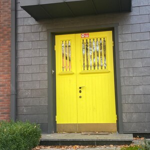May include: A bright yellow double door with vertical wooden slats and a black handle. The door is set in a gray stone wall with a dark gray trim. A small green bush and a stone walkway are in the foreground.