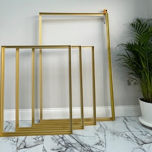 Puede incluir: Cuatro marcos de fotos de color dorado de diferentes tamaños están dispuestos en un suelo de mármol blanco. Los marcos están todos vacíos y tienen un diseño simple y moderno.