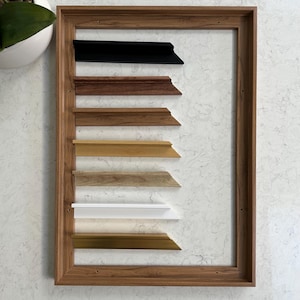 May include: A selection of picture frame mouldings are displayed within a wooden frame. The mouldings are in various colours and styles, including black, brown, gold, and white.