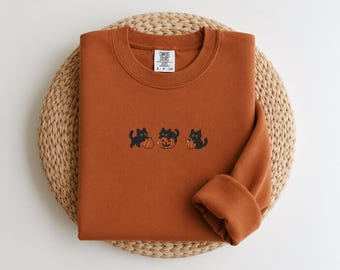May include: A rust-colored crewneck sweatshirt featuring an embroidered design of three black cats interacting with orange pumpkins. The interior neck label displays the text Comfort Colors, S-P-CH. The garment has long sleeves and a ribbed collar.
