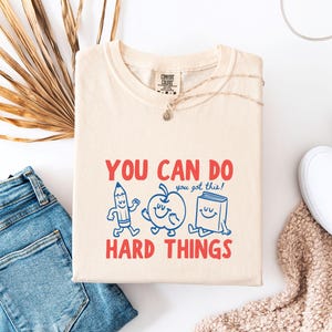May include: A cream-coloured t-shirt featuring a graphic with red text that reads YOU CAN DO HARD THINGS and blue line art of a smiling pencil, apple, and book with the text you got this! The shirt has a crew neckline and a Comfort Colors label.